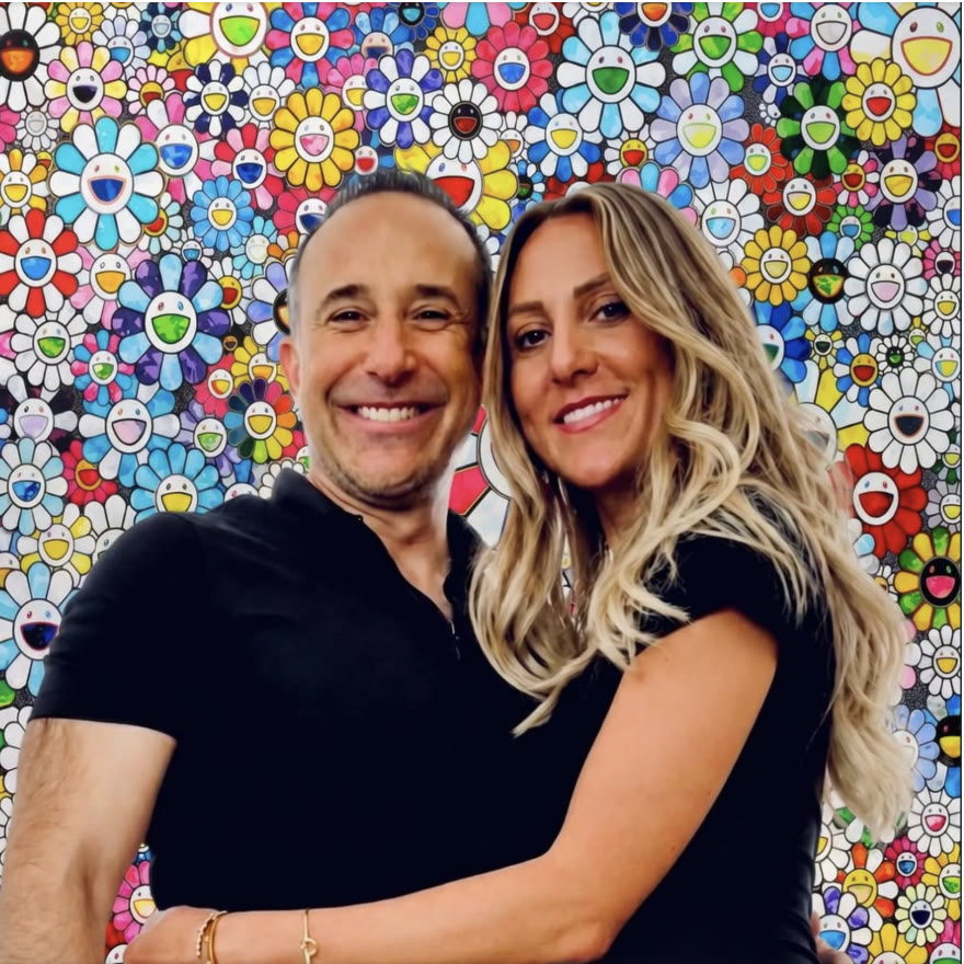 Robert and Cassie, arms around each other, in front of a wall of smiling Murakami flowers.